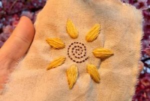 Hand holding brown square with sun-shaped embroidery.