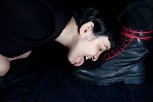 Woman dressed in black licking officer's black boot with red laces against a black background.