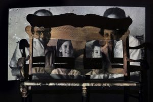 Black background with projection of father, mother, daughter and son behind bed frame.
